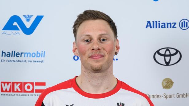 Stefan Egger-Riedmüller: Ein junger Mann im weißen Sporttrikot mit roten Akzenten steht vor einer Sponsorenwand und lächelt leicht.