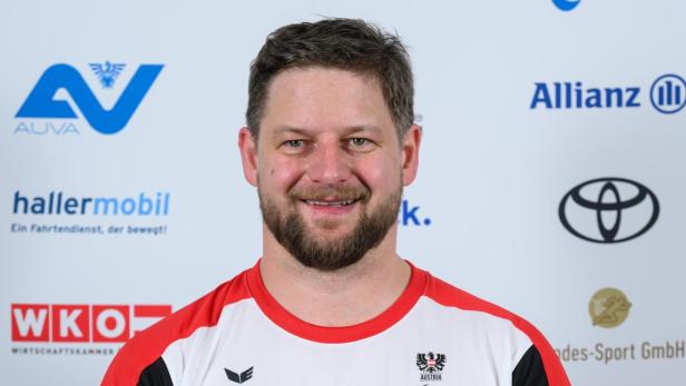 Michael Scharnagl: Ein Mann mit Bart trägt ein weißes Sportshirt mit roten Akzenten und steht vor einer Sponsorenwand.
