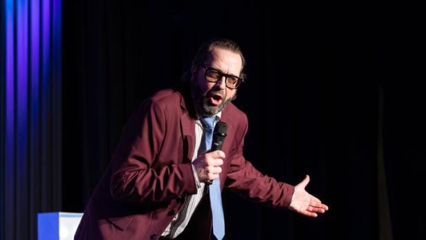Ein Mann im bordeauxfarbenen Anzug mit Mikrofon steht gestikulierend auf einer Bühne unter Scheinwerfern.