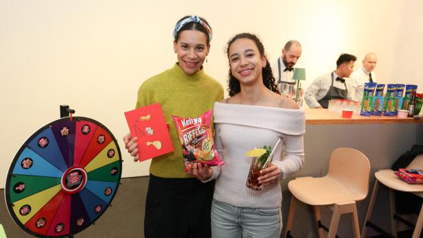 Zwei junge Frauen stehen lächelnd vor einer Bar, eine hält Chips und eine Karte, die andere ein Getränk mit Schirmchen.