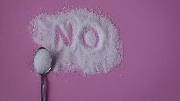 Ein Löffel Zucker liegt neben einem Haufen Zucker, in den das Wort „NO“ geschrieben ist, auf rosa Hintergrund.