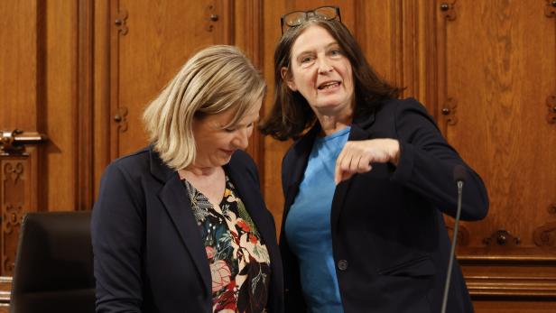 Zwei Frauen in Anzügen stehen vor einer hölzernen Wand und diskutieren.