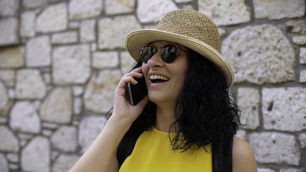 Frau mit Sonnenbrille und Strohhut lacht beim Telefonieren vor einer Steinmauer.