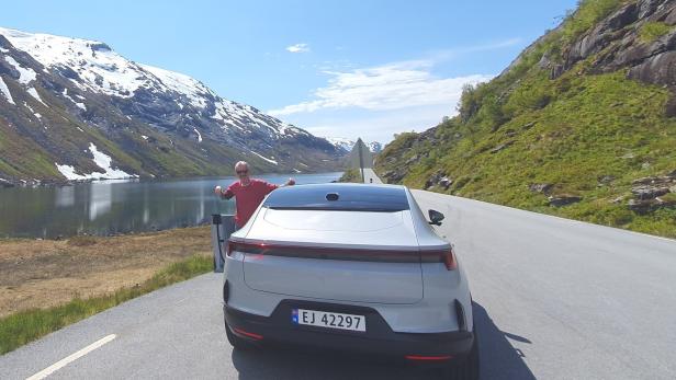 Ein Mann steht neben einem weißen Auto auf einer Straße an einem See, umgeben von Bergen mit Schnee und grüner Vegetation.