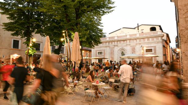 Ein belebter Platz mit vielen Menschen an Tischen unter Sonnenschirmen vor einem Gebäude.
