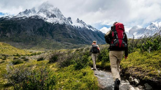Zwei Wanderer mit Rucksäcken gehen auf einem schmalen Bergpfad vor schneebedeckten Gipfeln durch eine alpine Landschaft.