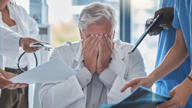 Ein älterer Arzt sitzt gestresst am Schreibtisch und hält sich das Gesicht, während mehrere Personen ihm Unterlagen und Geräte reichen.