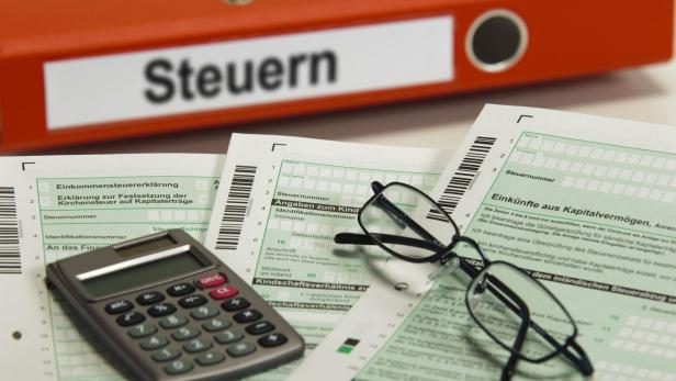 Auf Steuerformularen liegen eine Brille, ein Taschenrechner und ein Ordner mit der Aufschrift "Steuern".