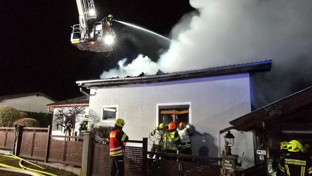 Mehrere Feuerwehrleute bekämpfen nachts einen Brand in einem Wohnhaus, dichter Rauch steigt vom Dach auf.