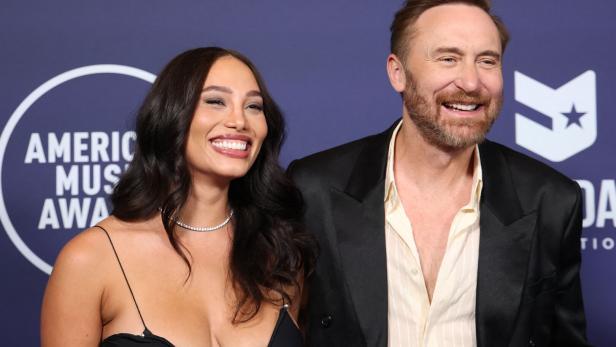 David Guetta und seine Freundin Jessica Ledon lächeln vor einem American Music Awards Hintergrund.