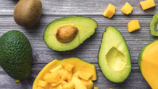 Geschnittene Avocado, Mango und Kiwi liegen zusammen mit ganzen Früchten auf einer Holzoberfläche.