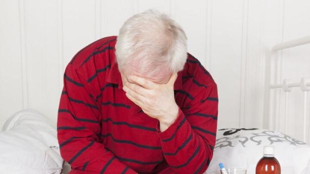 Älterer Mann im roten Schlafanzug sitzt erschöpft am Bettrand und hält den Kopf in der Hand, neben ihm Medikamente auf einem Tisch.