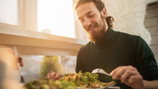 Ein Mann mit Bart sitzt am Tisch und isst bei Sonnenlicht einen Teller mit frischem Salat.