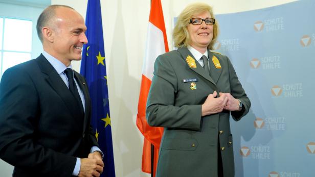 Ein Mann im Anzug und eine Frau in Uniform stehen vor Flaggen der EU und Österreichs.