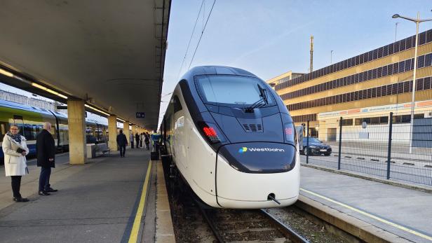 Moderner Westbahn-Zug steht am Bahnsteig, Menschen warten daneben.