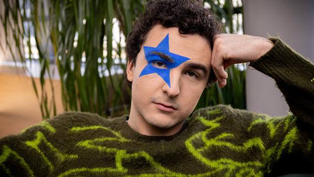 Eine Person mit lockigen Haaren trägt einen grünen Pullover und hat einen großen blauen Stern um das rechte Auge gemalt.