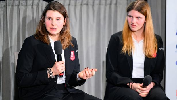 Zwei Frauen in Anzügen sitzen vor einem Vorhang, eine spricht ins Mikrofon.
