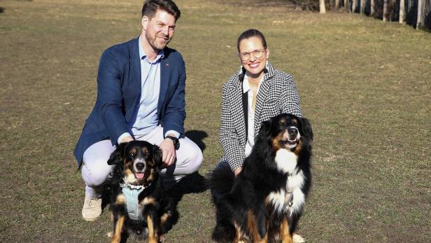 Ein Mann und eine Frau knien auf einer Wiese und lächeln, neben ihnen sitzen zwei große, schwarz-braune Hunde.