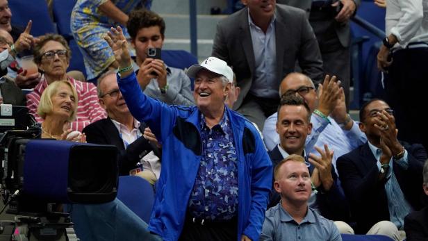 Ein älterer Mann in blauer Jacke und weißer Kappe winkt lächelnd in einer jubelnden Menschenmenge.