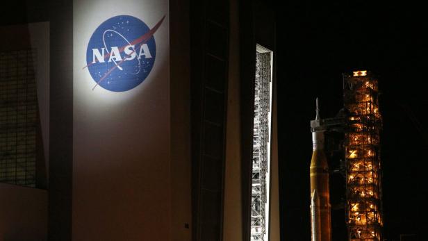 Ein großes NASA-Logo an einem Gebäude, daneben eine beleuchtete Rakete auf einer Startrampe bei Nacht.