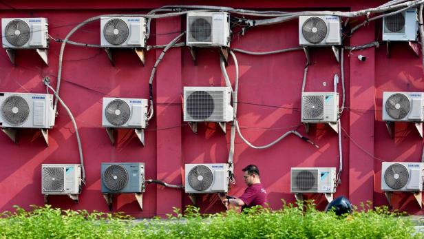 Mehrere Klimaanlagen sind an einer roten Wand montiert, davor geht ein Mann entlang grüner Büsche.