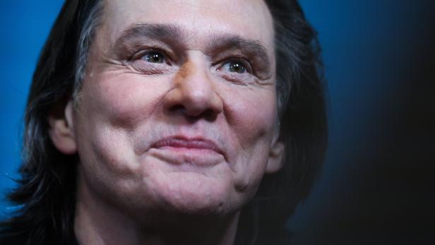 Jim Carrey lächelt mit leicht geneigtem Kopf vor einem blauen Hintergrund.