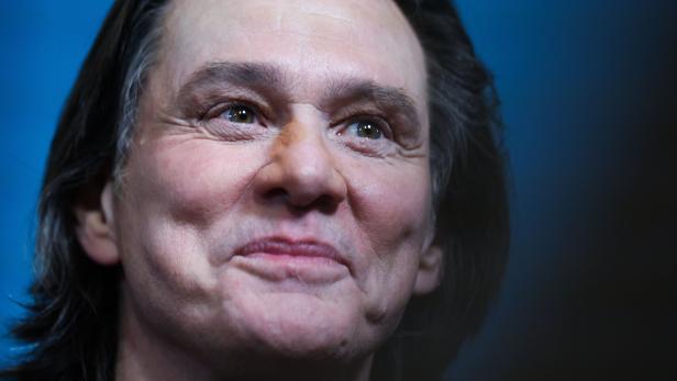 Eine Person mit mittellangen dunklen Haaren lächelt leicht vor blauem Hintergrund.