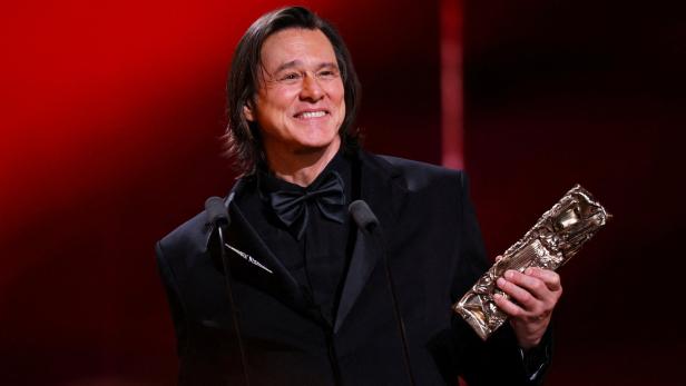 Jim Carreyim schwarzen Anzug mit Fliege hält eine goldene Trophäe und lächelt vor rotem Hintergrund.