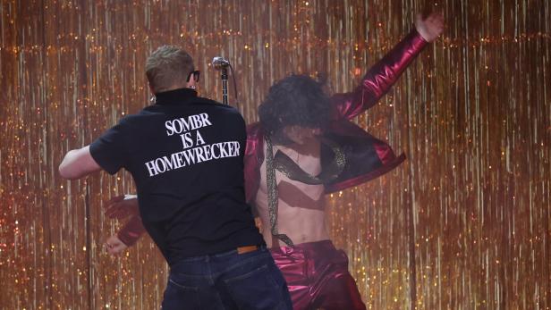 Eine Person im schwarzen T-Shirt mit weißem Aufdruck stört eine Bühnenperformance vor goldenem Lamettavorhang.