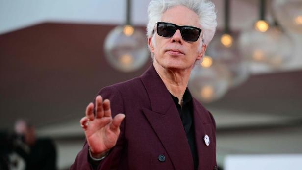 Jim Jarmusch mit Sonnenbrille und grauem Haar trägt einen bordeauxroten Anzug und winkt.