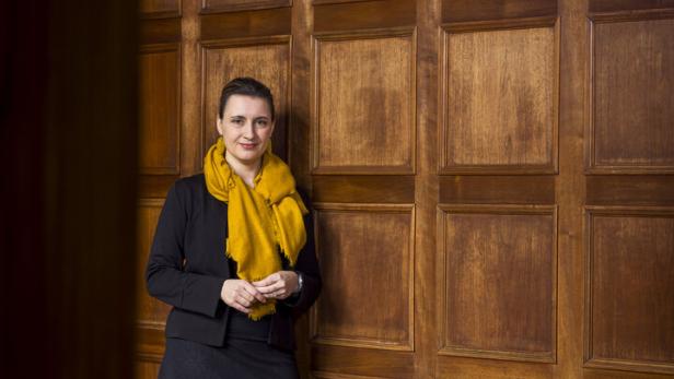 Eine Frau mit gelbem Schal lehnt an einer holzvertäfelten Wand und blickt freundlich in die Kamera.