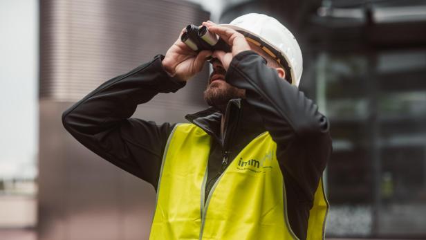 Ein Mann mit Schutzhelm und Warnweste blickt mit einem Fernglas nach oben.