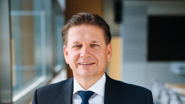 Ein Mann im dunklen Anzug und blauer Krawatte lächelt in einem hellen Büro.