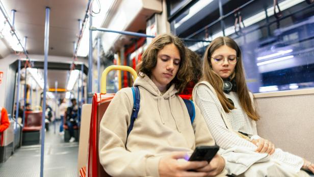 Zwei Jugendliche sitzen nebeneinander in einer Straßenbahn, einer schaut auf sein Handy, die andere blickt nach unten.