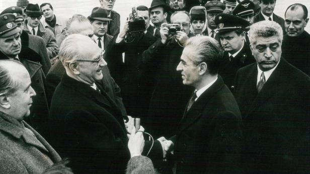Mehrere Männer in Anzügen und Uniformen begrüßen sich auf einem Flughafen, umgeben von Fotografen und Journalisten.