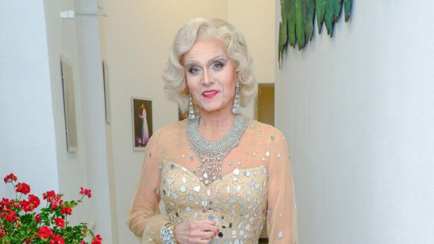 Eine elegant gekleidete Person mit blondem Haar trägt ein glitzerndes, beigefarbenes Abendkleid mit hohem Beinschlitz und auffälligem Schmuck.