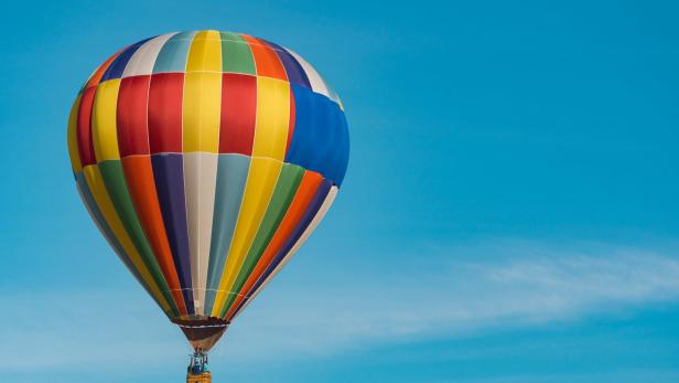 Ein bunter Heißluftballon mit zwei Personen im Korb schwebt vor blauem Himmel.