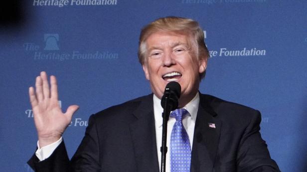 Donald Trump steht auf einer Bühne und hebt die rechte Hand.