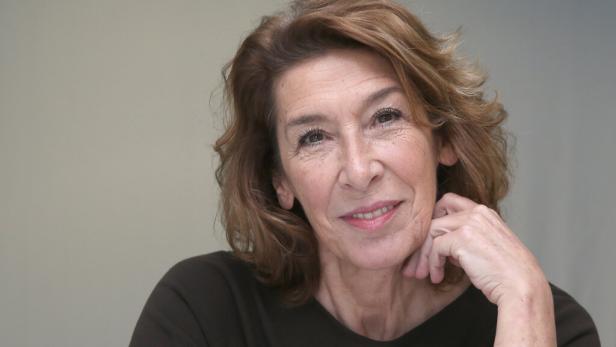 Adele Neuhauser mit braunem Haar und dunklem Oberteil lächelt, den Kopf auf ihre Hand gestützt.