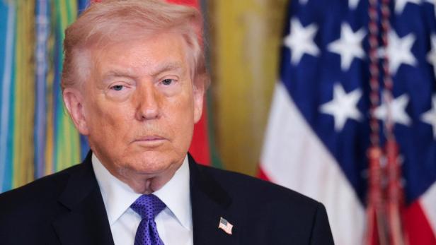 Donald Trump mit dunklem Anzug und gepunkteter Krawatte vor Flaggen.