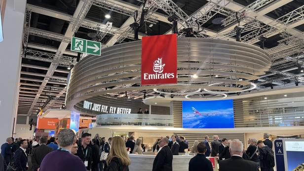 Viele Menschen stehen an einem modernen Messestand von Emirates mit großem Logo und Bildschirm, der ein Flugzeug zeigt.
