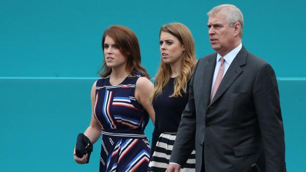 Prinz Andrew geht mit Prinzessin Beatrice und Prinzessin Eugenie vor einem türkisfarbenen Hintergrund.
