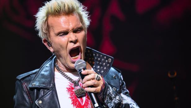 Billy Idol singt mit offenem Mund in ein Mikrofon, bekleidet mit Lederjacke und Kette.