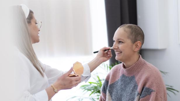 Eine Frau trägt einer lächelnden Person mit sehr kurzen Haaren Make-up mit einem Pinsel auf.
