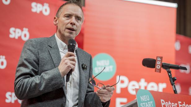 Ein Mann im grauen Sakko spricht an einem Rednerpult mit Mikrofon vor rotem Hintergrund mit SPÖ-Logos.