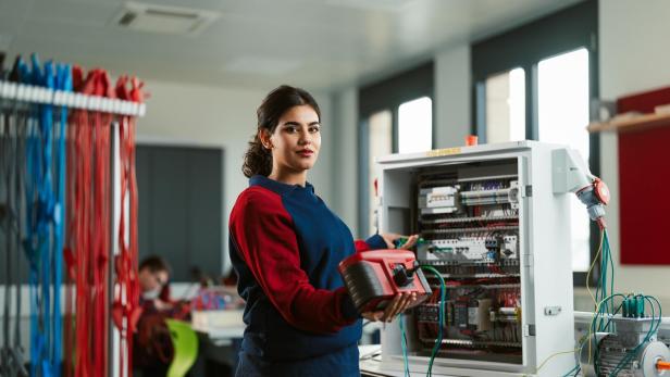 Eine junge Frau prüft einen offenen Schaltschrank mit einem Messgerät in einem hellen Raum.