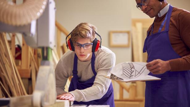 Ein junger Mann mit Schutzbrille arbeitet an einer Säge, während ein älterer Mann mit Bauplan zusieht.