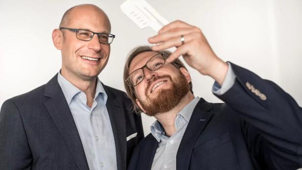 Zwei Männer in Anzügen betrachten lächelnd einen weißen Gegenstand vor einem hellen Hintergrund.