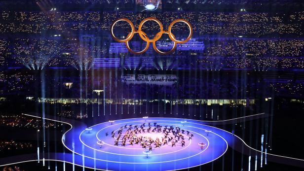 Eröffnungszeremonie mit vielen Menschen auf der Bühne und den olympischen Ringen im Hintergrund .