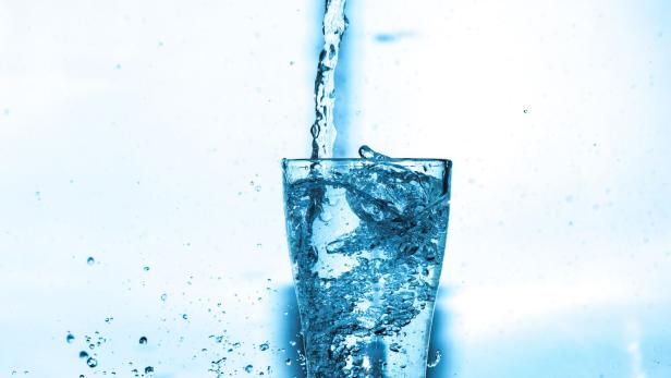 Ein Glas wird mit spritzendem Wasser gefüllt, während Tropfen und Wellen um das Glas entstehen.